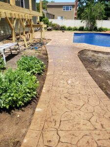 Stamped concrete walkway curves around a garden and leads to a backyard pool area beside a wooden structure.