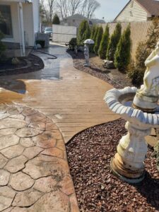 A backyard walkway with stamped concrete, a wooden path, a white statue, and a decorative birdbath fountain.