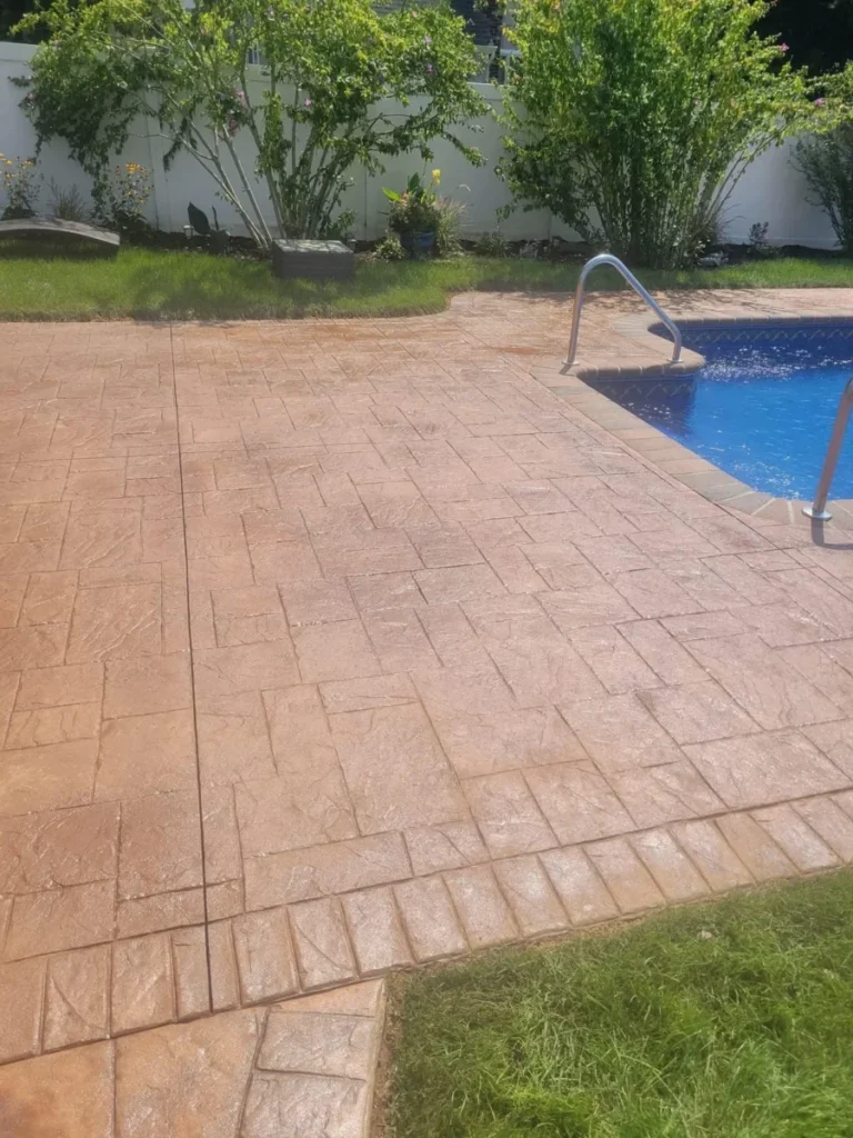 Stamped concrete patio next to a swimming pool with metal handrails, bordered by grass and shrubs.