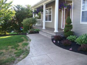 Concrete walkway leads to a porch with columns, hanging plants, and landscaped garden beds with small American flags.