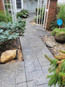 Gray stamped concrete walkway with stone accents leads to a house entrance, bordered by plants and a metal gate.