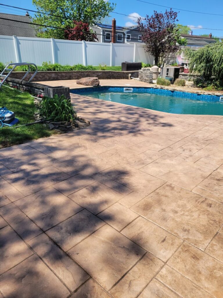 A backyard with a stamped concrete patio, in-ground pool, and landscaped plants on a sunny day.