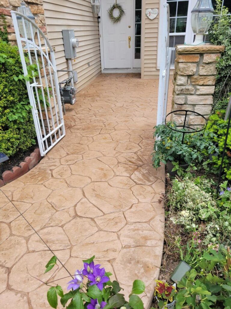 A stamped concrete walkway leads to a white door, bordered by plants and a metal gate on the left.