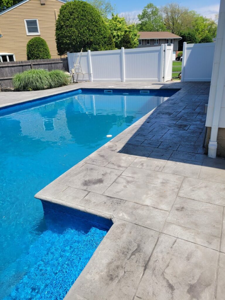 Rectangular backyard swimming pool with a concrete deck, surrounded by fences and greenery on a sunny day.