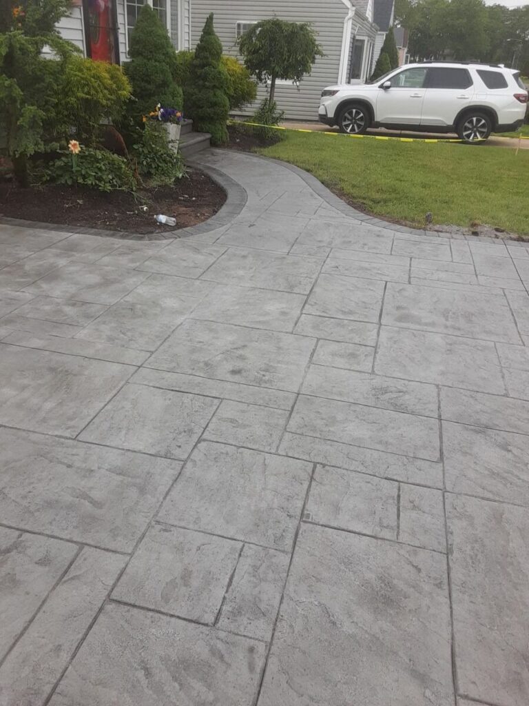 Stamped concrete driveway and walkway lead to a house with landscaping and a white SUV parked on the street.