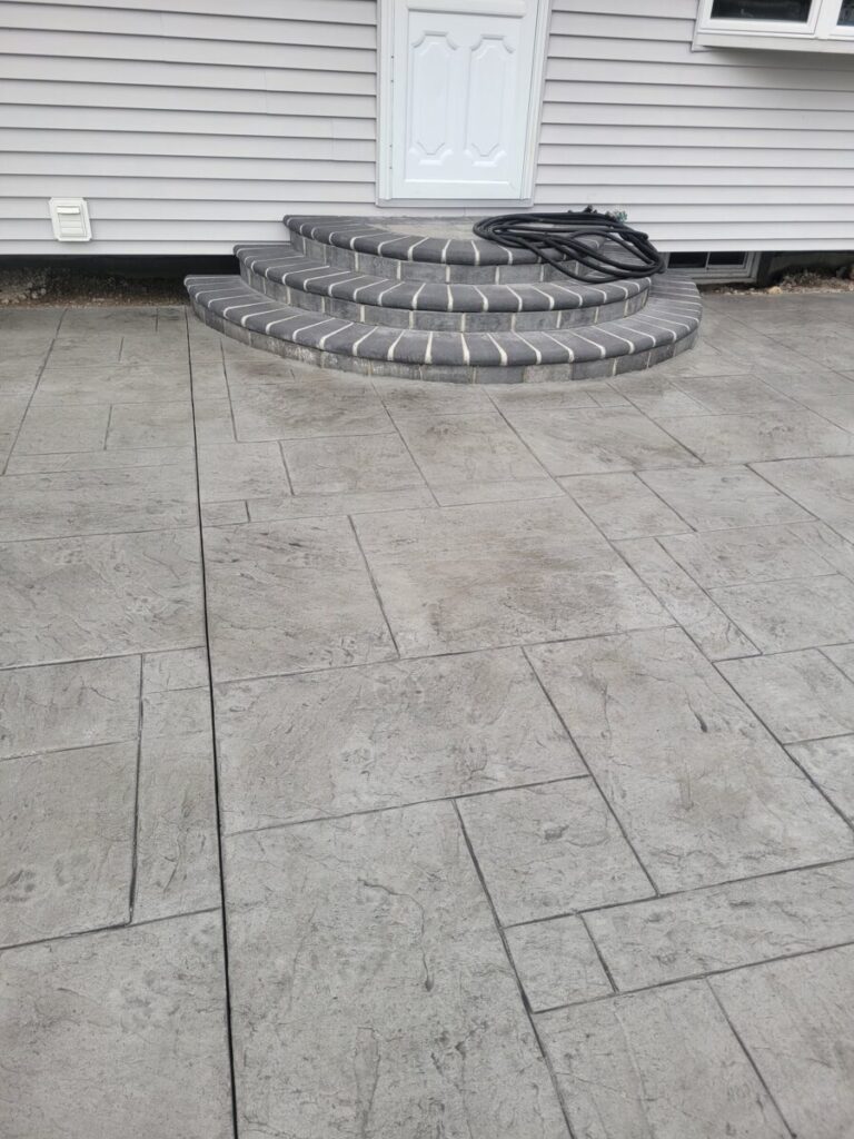 Concrete patio with rectangular patterns leads to three semi-circular gray steps at a white door of a house.