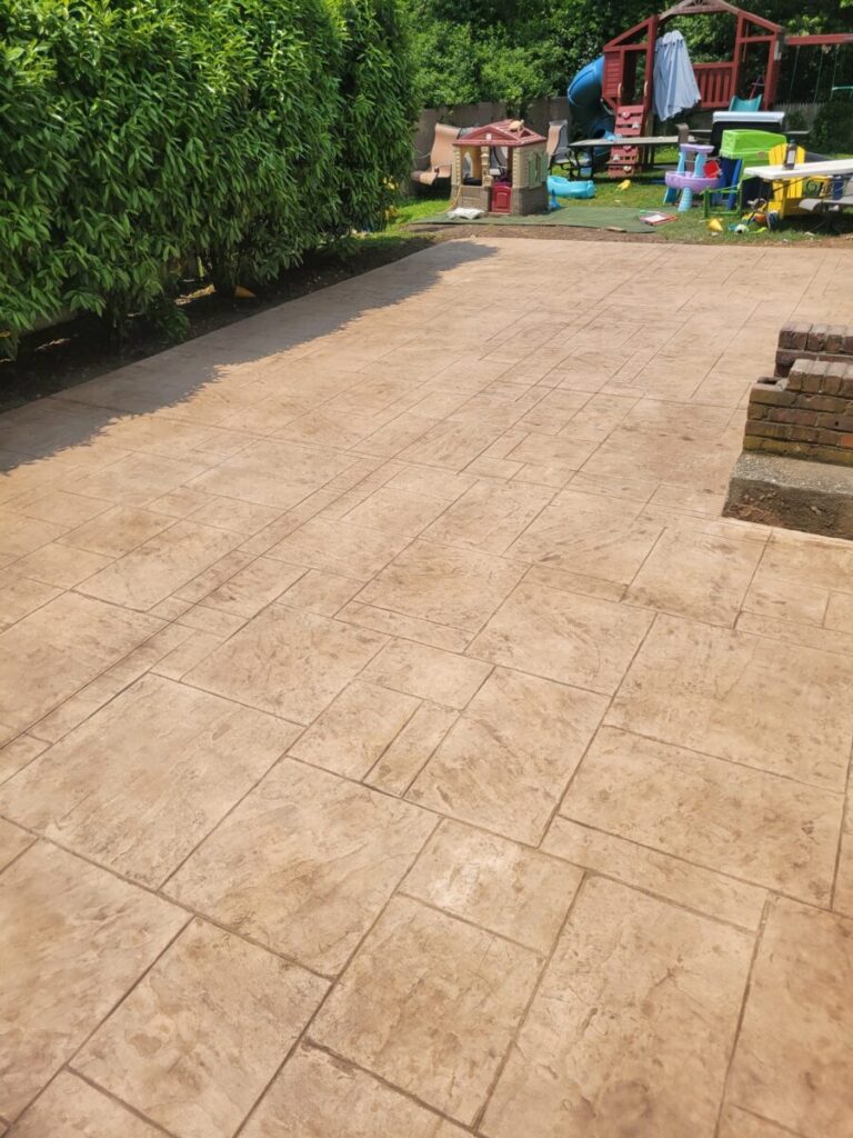 Stamped concrete patio with a hedge on the left and children's play equipment in the background.