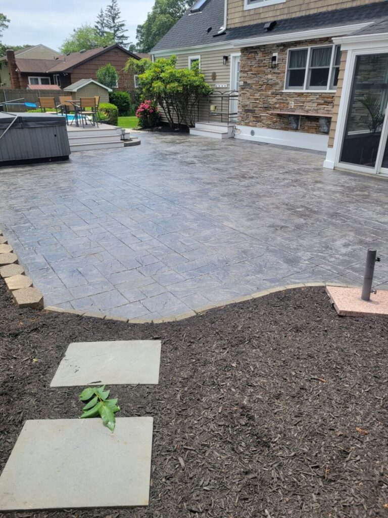 A stamped concrete patio beside a house with landscaped mulch, stepping stones, and a hot tub in the corner.