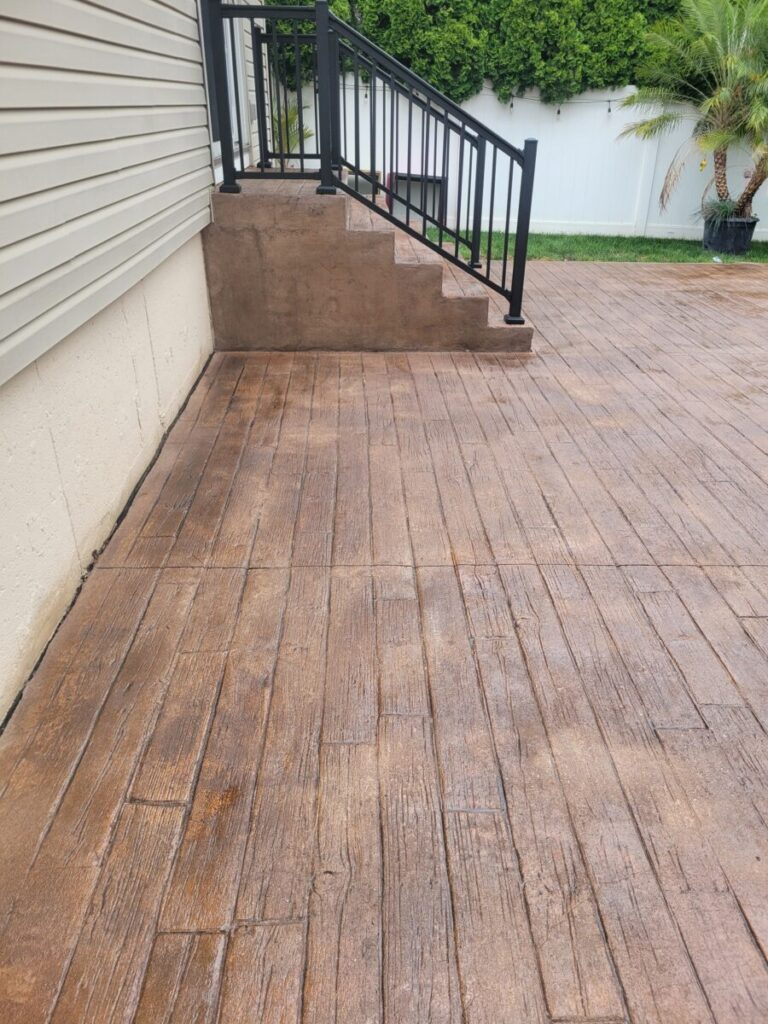 Stamped concrete patio with a wood plank pattern, leading to stairs with a black metal railing and greenery in the background.