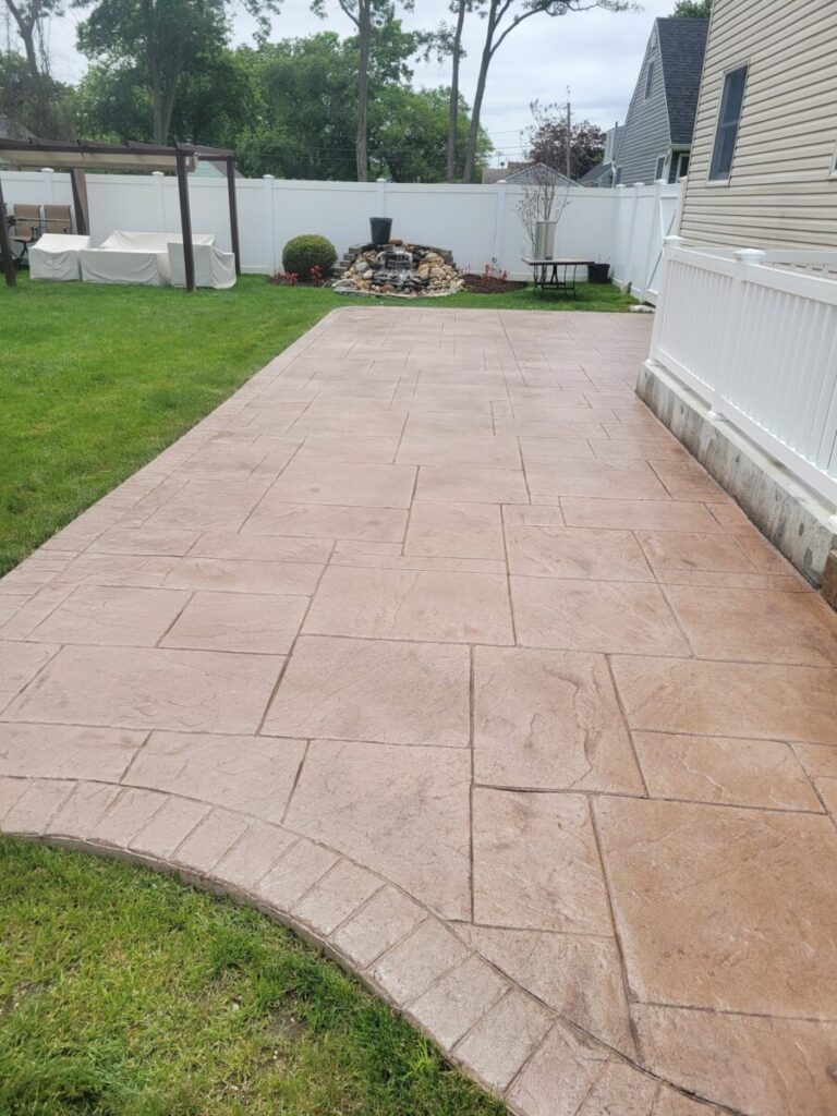 A stamped concrete patio extends from a house toward a small rock waterfall and landscaped area in a fenced backyard.
