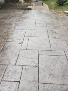 Outdoor area with light gray, stamped concrete in a rectangular pattern, adjacent to grass and patio furniture.