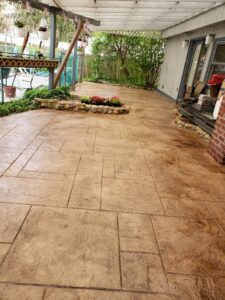 Spacious patio with stamped concrete flooring, a small raised flower bed, and plants along the edges near a house.