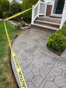 Yellow caution tape blocks off a newly paved walkway leading up to steps beside a garden and shrubs.