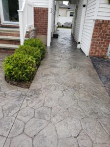 A stamped concrete walkway runs between a house and a hedge, leading to a backyard seating area.
