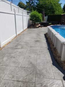 Concrete walkway beside an above-ground pool, bordered by a white fence and gravel, with trees in the background.
