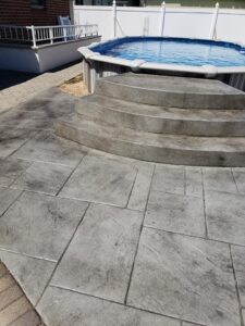 Outdoor above-ground pool with curved concrete steps and stamped concrete patio surface in sunlight.