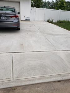 A gray car is parked on a concrete driveway with circular marks in front of a white garage and fence.