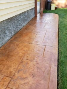 Stamped concrete patio with a textured, brown finish next to a house with yellow siding and a grassy lawn.
